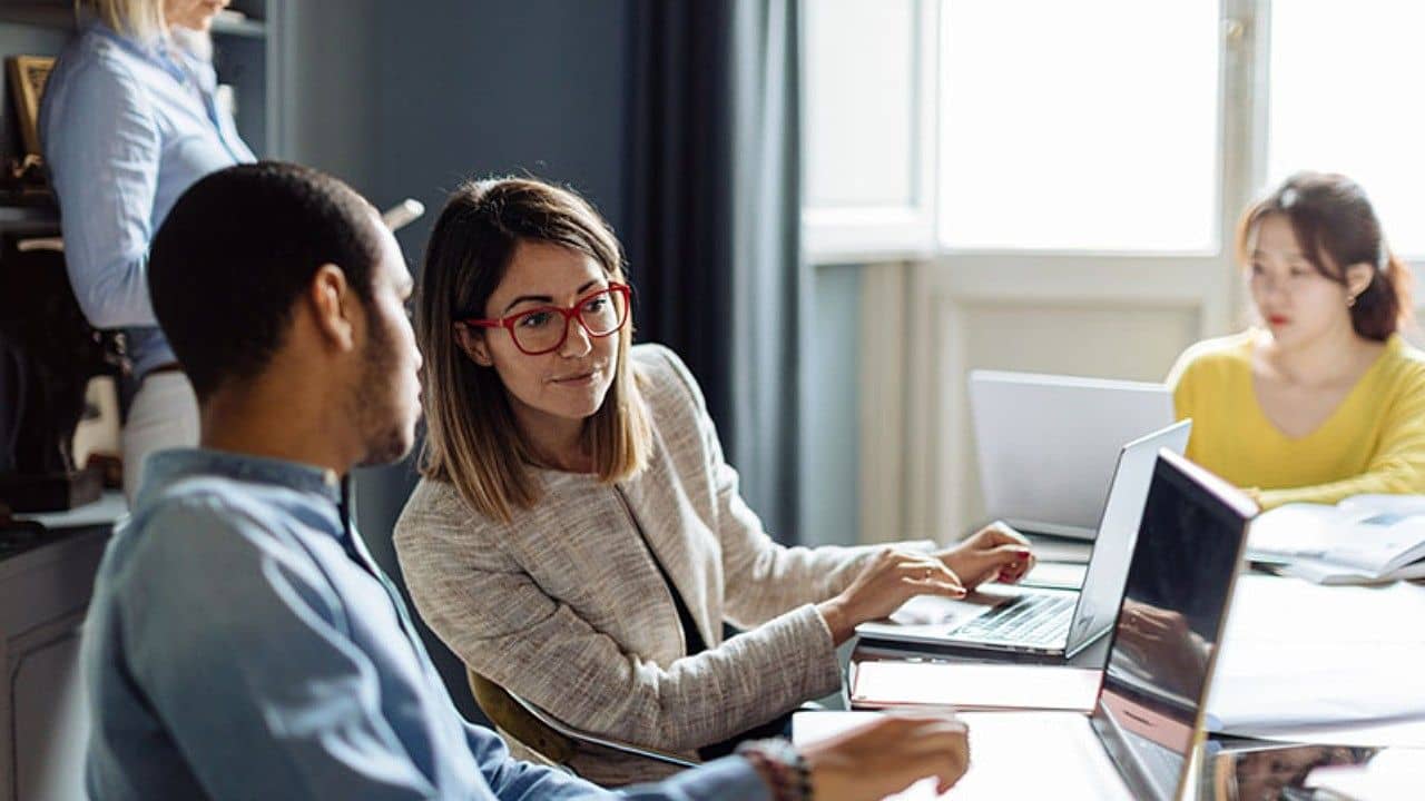 Three people work at laptops around a table in a bright office, with a fourth person standing in the background. One woman with glasses is talking to a man seated beside her.