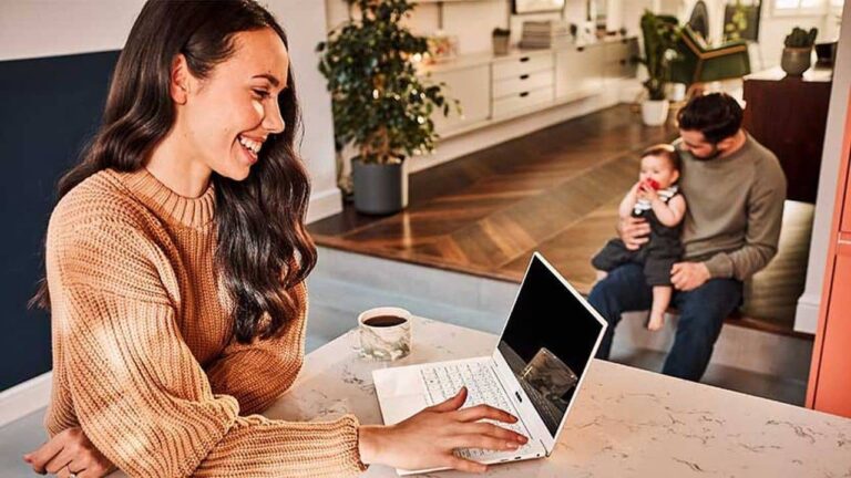 A woman sits at a kitchen counter using a laptop and smiling, with a cup of coffee nearby. In the background, a man sits on steps holding a baby in a modern, well-lit home.