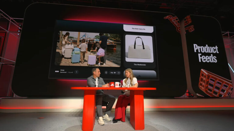 Two presenters sit at a red table on stage discussing product feeds, with a large screen behind them displaying luggage products titled "The Kids Carry-On" and "The Weekender" alongside a video of people with suitcases at a waterfront location.