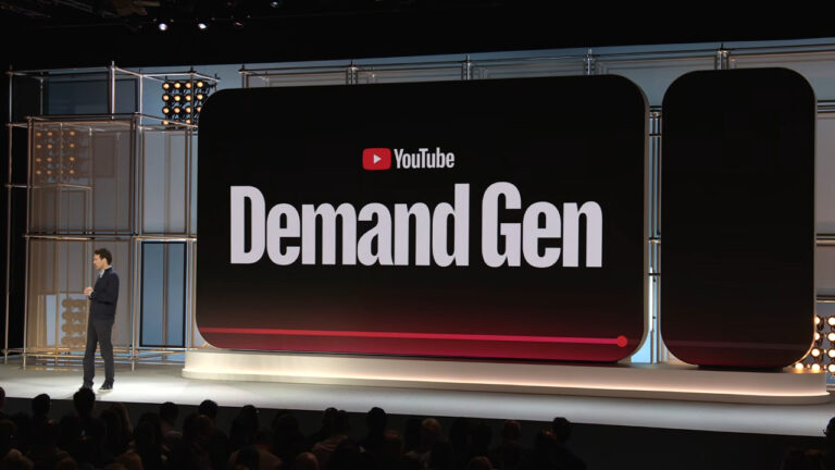 A presenter stands on stage next to a large display screen showing the YouTube logo and "Demand Gen" text in white letters against a black background with red accent lighting.