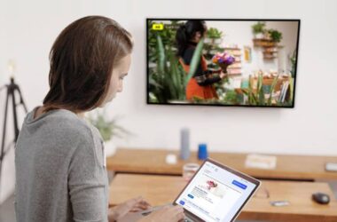 A woman using a laptop with a flower shop website on the screen, sitting in a modern living room. The television displays CTV content showing someone holding a bouquet of flowers, while she explores Microsoft Ads for LinkedIn Profile Targeting.