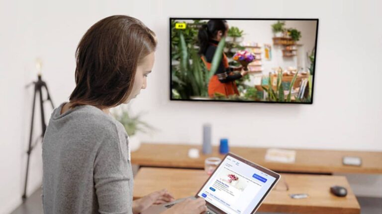 A woman using a laptop with a flower shop website on the screen, sitting in a modern living room. The television displays CTV content showing someone holding a bouquet of flowers, while she explores Microsoft Ads for LinkedIn Profile Targeting.