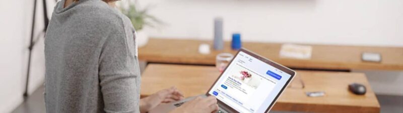 A woman using a laptop with a flower shop website on the screen, sitting in a modern living room. The television displays CTV content showing someone holding a bouquet of flowers, while she explores Microsoft Ads for LinkedIn Profile Targeting.