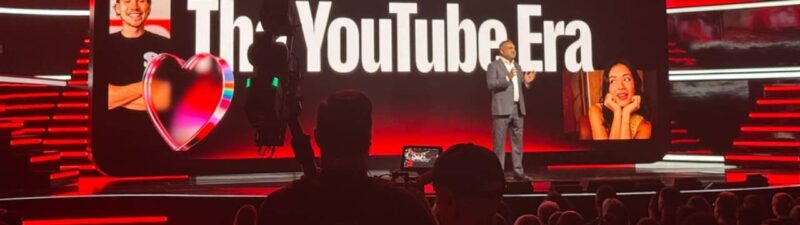 A presenter stands on stage in front of a large screen displaying "Welcome to The YouTube Era" with the YouTube logo, surrounded by neon red lighting and images of content creators on a dark background.