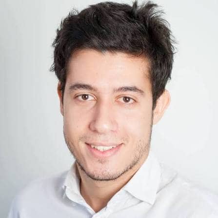 A young man with short, dark, slightly messy hair and light facial hair is smiling at the camera. He is wearing a white collared shirt and is posed against a plain, light background.