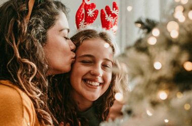 Two people smiling near a decorated Christmas tree, one wearing antlers and being kissed on the cheek by the other, with warm festive lights in the foreground.