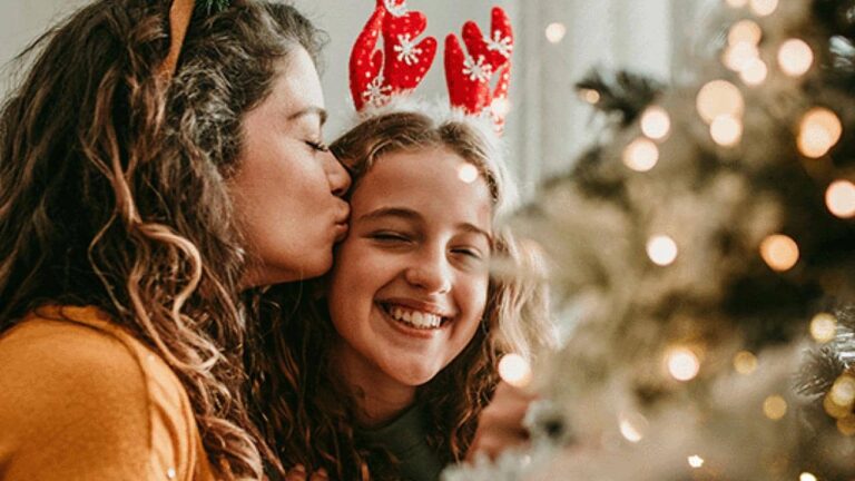 Two people smiling near a decorated Christmas tree, one wearing antlers and being kissed on the cheek by the other, with warm festive lights in the foreground.